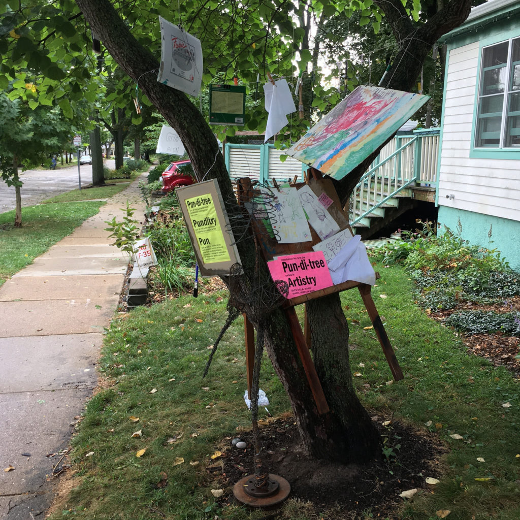 Tree with drawings, paintings, art supplies handing from it, plus an easel perched between two branches